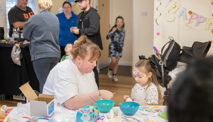 A woman in a white tshirt sits with a little girl with her face painted at a table. They are making something together. On the table in front of them are two blue plastic bowls with paintbrushes in them, and other craft supplies. There are other adults and two children blurred in the background. It looks like a community centre. The word "Happy" is strung up in the background.