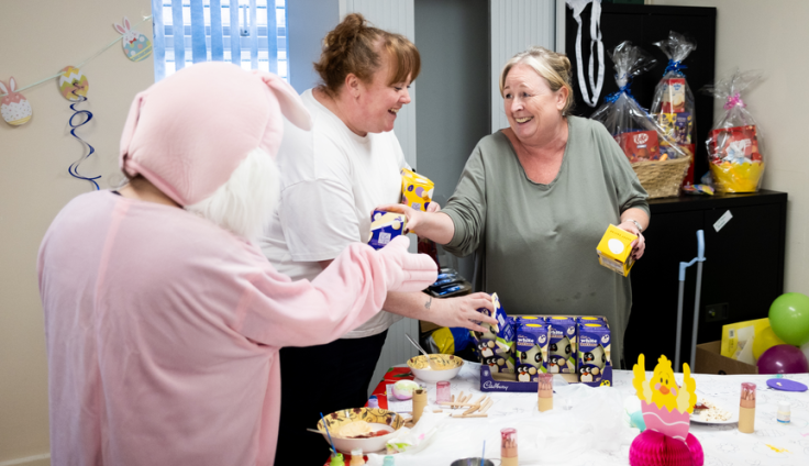 Two women laugh as they pick up boxes of chocolate Easter eggs off a table and give them to someone dressed in a pink bunny suit. There is bunting with Easter eggs strung up against the wall behind them and baskets of wrapped chocolate boxes on a black sideboard. On the table in front of them are pencils, an Easter chick decoration, and balloons.