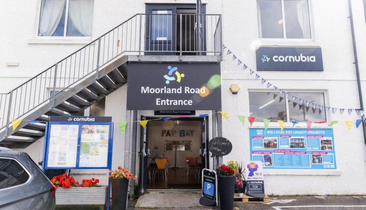 A ground floor entrance to the Cornubia Centre, with a sign above the door reading "Moorland Road Entrance". There is a community notice board on the left side of the door, and a large bright blue information board to the right, showcasing Par Bay Big Local key legacy projects.
