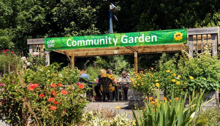 A group of people sitting around a table doing crafts under a pergola on a sunny day in the Par Bay Community Garden.
