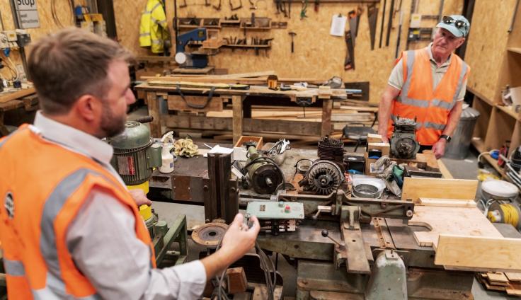Two men wearing orange hi-vis vests, working in a tool shed.