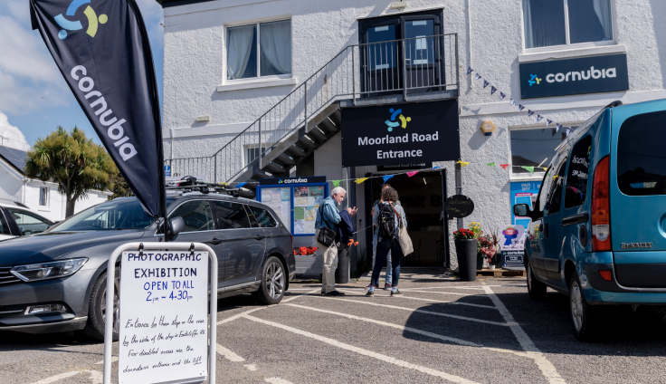 A white washed two-storey building has a sign on it reading 'Cornubia'. Three people stand outside the entrance in the car park. A grey car is parked on the left, and a blue on the right. There is also a sign in the foreground that reads 'photographic exhibition, open to all'. There are red flowers in pots outside the entrance to the building on the ground floor. A set of steps leads to some black double doors on the first floor. On the left, a tree, a white house and blue sky are visible.