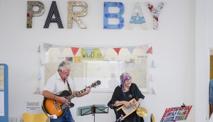 An elderly man sings and plays guitar while an elderly woman next to him with glasses and grey and pink hair plays a washboard and holds a cymbal. There is a noticeboard behind them with bunting around it. Above that, giant letters covered in different designs are displayed on the wall, spelling out PAR BAY.