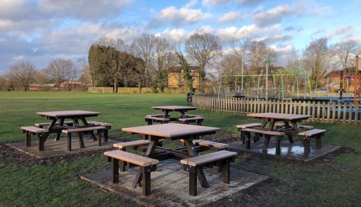 Four wooden picnic benches with seating in a grassy parkland.