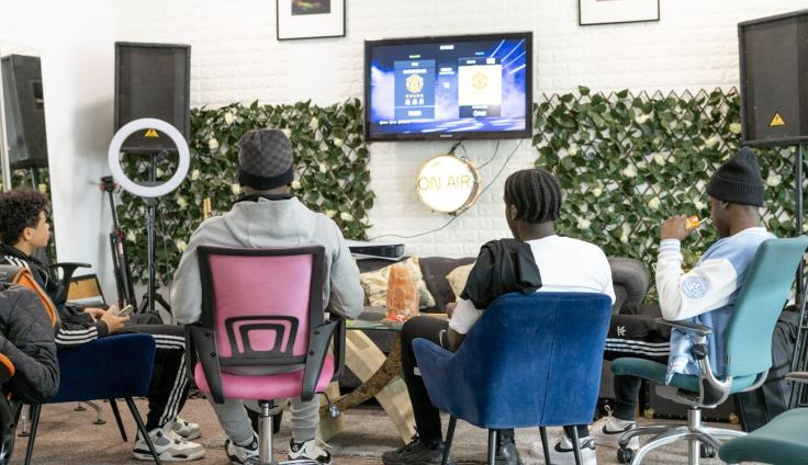 Four people in a room with chairs, facing a monitor, surrounded by tall sound speakers.
