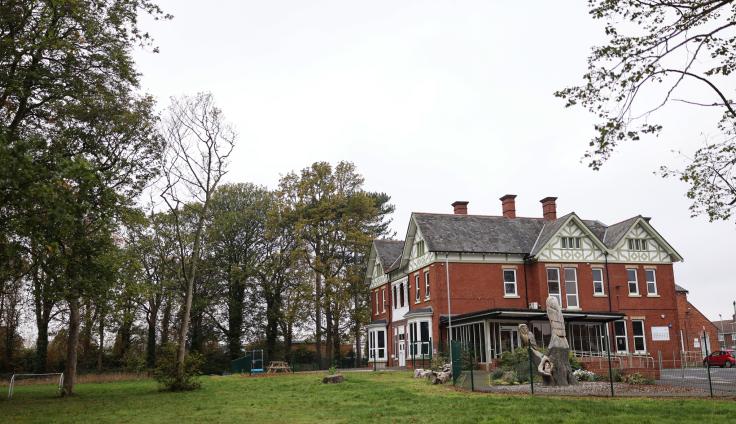 A large red-brick house set in a woodland area with grass to the front.