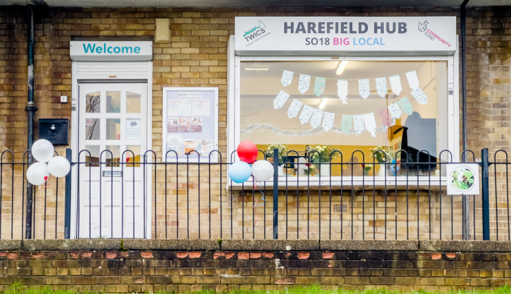 The front of Harefield Hub, a community centre housed in a yellow brick building. A large white sign above the main entrance reads "HAREFIELD HUB SO18 BIG LOCAL" with logos for TVICS and Big Community. To the left is a smaller "Welcome" sign above a notice board with various posted information. The building features large windows decorated with colorful bunting hanging inside. A black metal railing runs along the front of the building, adorned with white, red, and blue balloons suggesting a celebratory event.