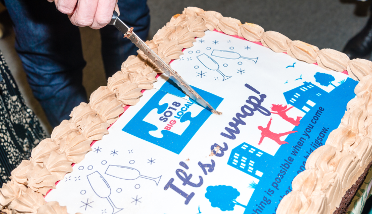 A hand holds a knife to cut into a large rectangular celebration cake with pale orange icing piped around the border. The white fondant top is decorated with blue and pink design elements including building silhouettes, trees, the silhouettes of two people jumping, birds and champagne flutes with sparkles around them. At the centre is the "SO18 BIG LOCAL" logo and purple text reads "It's a wrap!". The phrase "It is possible when you come together" appears in smaller text.