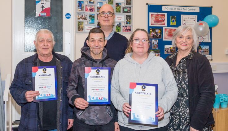 Five people stand together indoors holding certificates at a community recognition ceremony. From left to right: an older man in a dark checked shirt and cardigan, a man in a camouflage hoodie, a tall man with glasses and bald head wearing a dark sweater (standing in the back), a woman in a gray hoodie and glasses, and an older woman with short pink-silver hair wearing a dark top with a patterned scarf. The first three people hold blue and white certificates with "SO18 BIG LOCAL" branding. Behind them are bulletin boards displaying colourful photos and information, including one labeled "SO18BL in West End Carnival" with balloons attached.