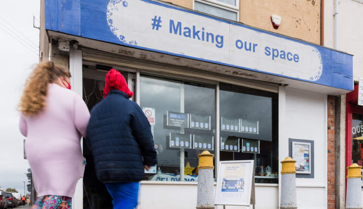 Two people walk towards the community hub on the high street. The person on the left is wearing a long pink cardigan and a red face mask, the person on the right has a red hood and a black jacket.