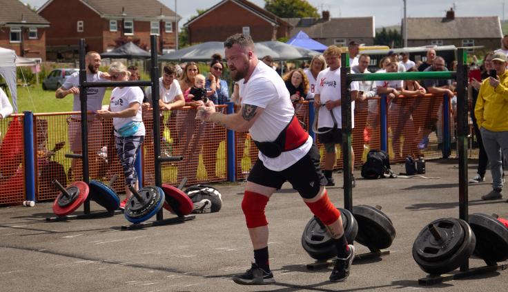 A man pulls wearing a weightlifting belt strides towards an obstacle out of shot. Weights and bars can be seen in a park, with an audience looking on, leaning over a barrier. Residential houses are in the background.