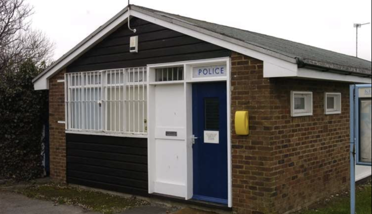 A small brick and shiplap building with an apex roof. It has a white door, the word "Police" over a blue door, and barred windows at the front. Two small windows can be seen on the side of the building.