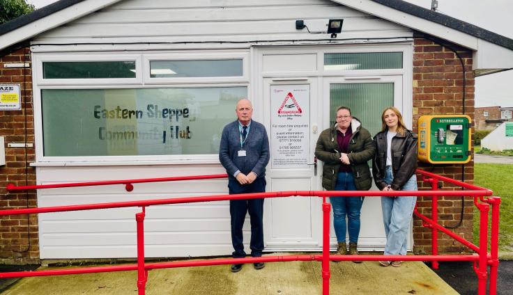 A man and two women stand outside a small brick and shiplap community building facing the camera. The window on the front of the building features frosted glass with the text 'Eastern Sheppey Community Hub' engraved on it. A red railing and accessible ramp lead up to the front door.
