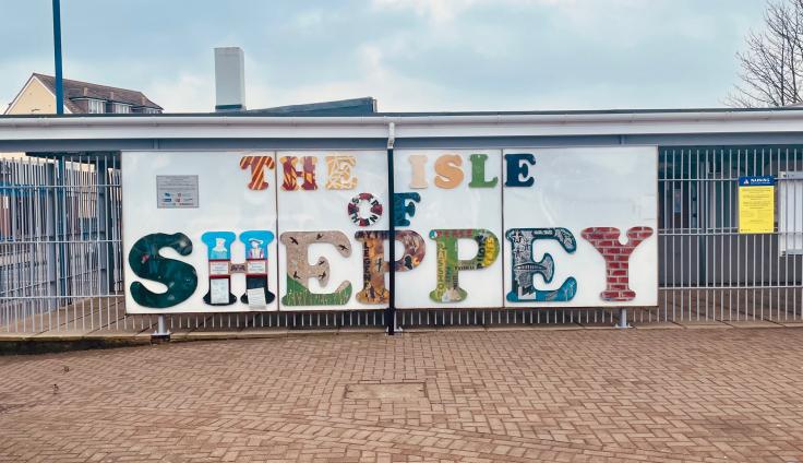 A sign attached to railings reads "The isle of sheppey". Each letter is a different pattern, colour or drawing. There is brick paving in the foreground and a cloudy blue sky in the background.
