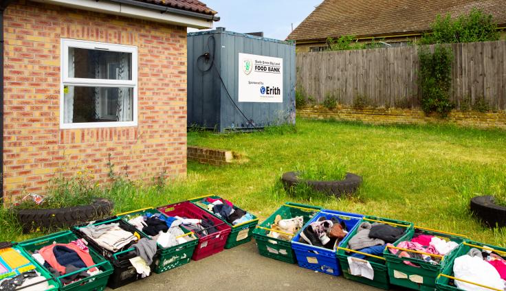 One-storey brick building with eleven green shopping crates full of clothes outside and a grey shipping container with a sign reading 'Slade Green Big Local Food Bank'.