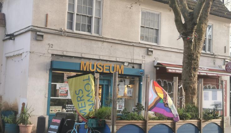 A white building with a blue sign reading 'museum', a seating area and flags outside.
