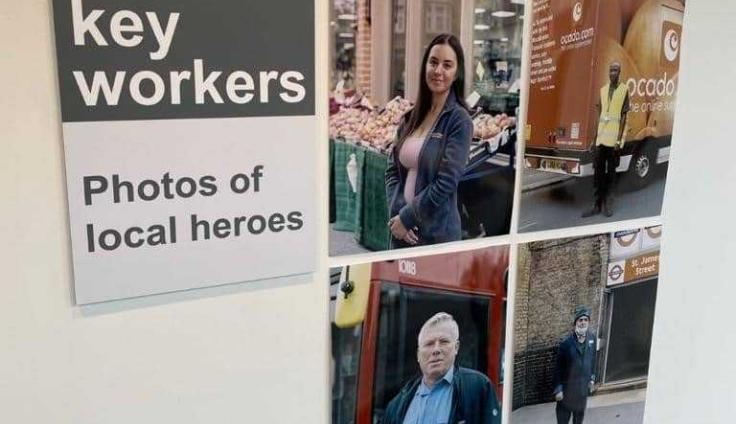 A close-up of a photographic display showing four local key workers, including: a young woman with long brown hair, a pink top and a blue jacket standing in front of a stall with onions on it; a young man in a high-vis vest standing in front of an orange food delivery van; an older man in a blue shirt and navy jacket standing in front of a red bus; and an older man with a white beard and glasses and a navy suit standing in front of the St James Street underground entrance.