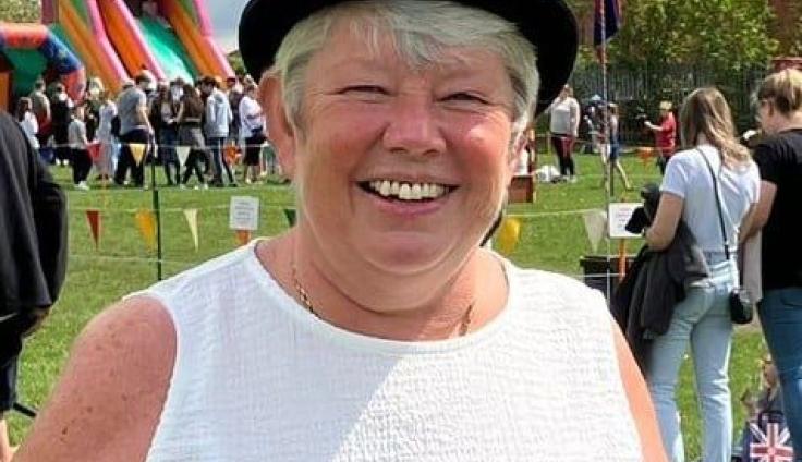 A woman wearing a black bowler hat, white tank top and a blue rosette, smiles at the camera. There is a bouncy castle in the background, bunting and crowds of people standing in a field.