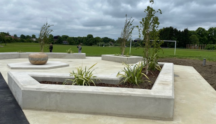A newly constructed concrete plaza featuring geometric white concrete planters and seating areas. The raised rectangular planters contain dark soil with newly planted young trees and grass-like plants. A large decorative stone bowl sits on one of the concrete platforms. The modern, minimalist design uses clean lines and right angles throughout the hardscaping. In the background, people can be seen walking across a large green field with football goalposts visible, and mature trees line the horizon under an overcast sky.