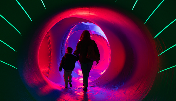 A woman and a child holding hands are silhouetted while walking through a light tunnel. The  circular tunnel has red, pink, blue lights around it. Bright green stripes of light radiated out of a dark background.