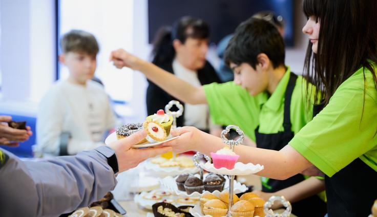 Children in aprons serving people plates of cakes from cake stands.