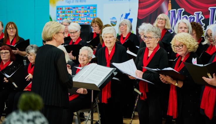 A group of women in black and red outfits standing with music folders open in front of a choir leader.