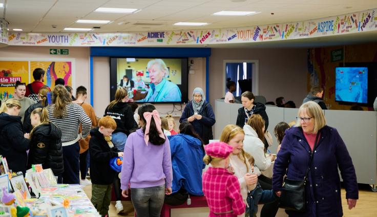 A gathering of people in a large room, watching a television screen next to tables with activities on them.