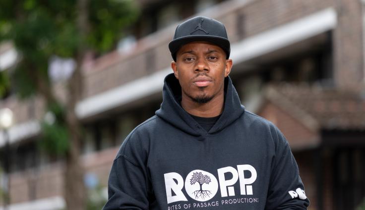 A young black man wearing a black cap and a black hoodie with ROPP on it looks straight at the camera. There is a blurred council estate building and a tree behind him.