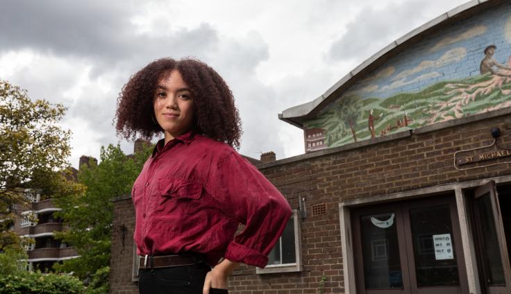 A young woman with curly hair smiles and looks straight at the camera, wearing a red shirt and black trousers. She stands in front of a brick building, an overcast sky and trees in the background.