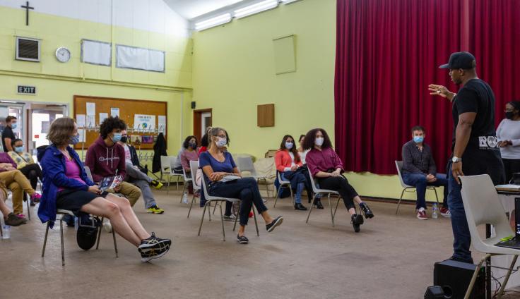 A diverse group of people wearing face masks sit in plastic chairs arranged in a socially distanced semicircle in a church hall. The room has pale yellow walls, red curtains on the right side, and institutional lighting. A man in a black t-shirt and baseball cap stands at the front, leading a workshop. The attendees, who include people of various ages and ethnicities, are listening attentively.