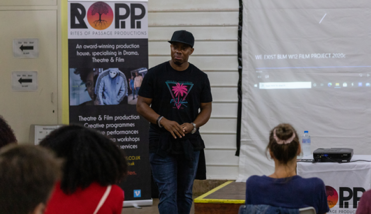 A man in a black t-shirt and cap standing beside a large banner for "ROPP - Rites of Passage Productions," describing it as an award-winning production house specializing in drama, theatre and film. Three people are sitting facing towards the man.