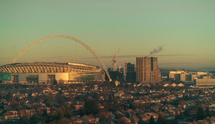 A landscape photo of Wembley at sunset. The Wembley Arena can be seen on the left of the horizon, with high rise buildings, cranes and lower buildings on the right. In the foreground are rows and rows of houses and trees.