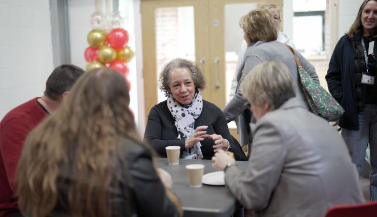 A group of people sit around a table listening to an older woman with curly short hair talking. She is gesturing and wears a polka dot white scarf and black jacket. There are two paper cups on the table and a couple of people standing up behind them by some wooden double doors. A woman is walking out. There are also red and gold balloons stringed up behind the group.