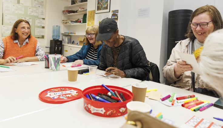 Four middle-aged women laughing while sitting around plastic trestle tables. There are colourful pens and bingo sheets on the table.