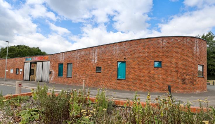 A low-angle, outdoor shot captures the exterior of a modern, curved brick community center under a bright, partly cloudy sky. The building is constructed of reddish-brown bricks and features a distinctive curved design. Several rectangular windows with teal-colored frames and blinds are visible. The main entrance, located to the left, consists of light-colored wooden double doors.