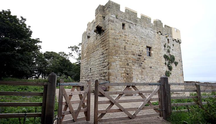 A turreted building rises up behind a wooden fence. Trees surround the old tower and there is a brick wall in the distance.