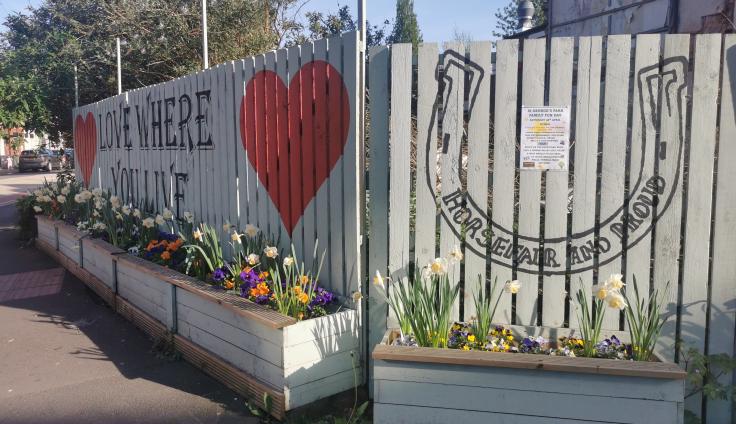 Painted fence panels with a horseshoe, red hearts and writing reading 'Love where you live' surrounded by colourful flower boxes