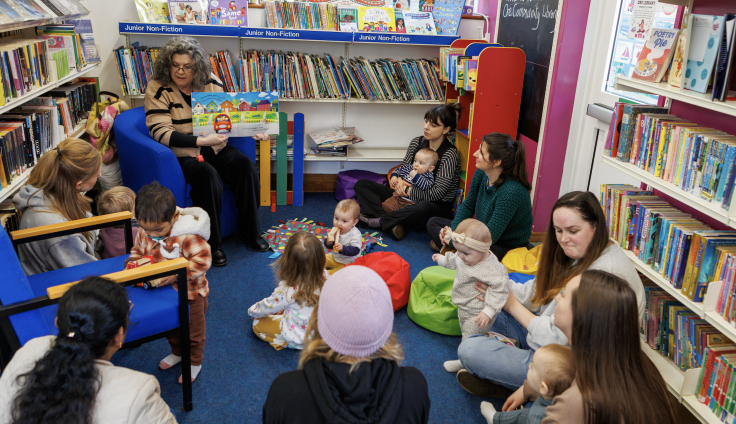 A library storytime session in progress in the children's section of a library. An older woman with curly grey hair and glasses sits in a blue chair reading a colourful picture book to a group of young children and their caregivers. The setting is surrounded by tall bookshelves filled with children's books, with blue shelf labels reading 'Junior Non-Fiction' visible above.