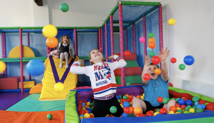 A girl and a boy play gleefully in a ball pit, throwing colourful balls up in the air. Behind them is a soft-play obstacle course, and a smiling girl coming down a yellow slide.