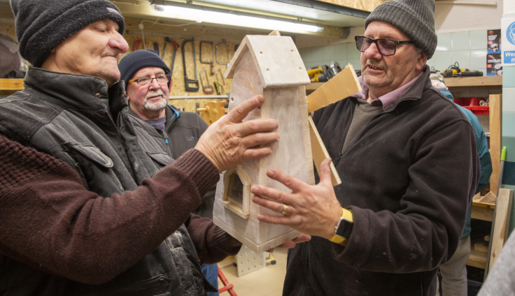 Three older men wearing woolen black hats and brown and black fleeces hold a wooden windmill together. Two of them have glasses. Woodworking tools and saws are hanging on a chipboard wall in the background and there is a strip light above them in the workshop.