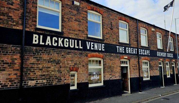 Brick building with a black painted band across the wall displaying large white text that reads “BLACKGULL VENUE THE GREAT ESCAPE GRIMSBY DOCKS.” The building has several windows and doors along the street, and a flag flying from a pole.