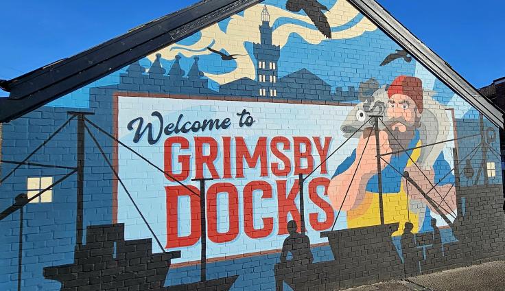 A painted mural on the side of a brick building depicts Grimsby Docks, with a painted sign across the middle that reads 'Welcome to Grimsby Docks' and a fisherman character with a fish around his neck.
