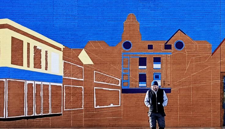A man wearing a hat and dark jacket walks in front of a large brick wall mural painted with simplified outlines of buildings against a bright blue background.