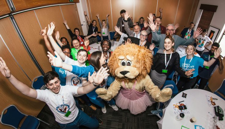 A group of young people with their hands in the air and a person wearing a lion costume and a tutu.