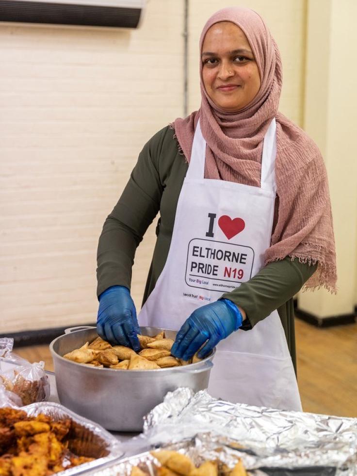 A woman serving food wearing a pink hijab and blue catering gloves, smiling at the camera.