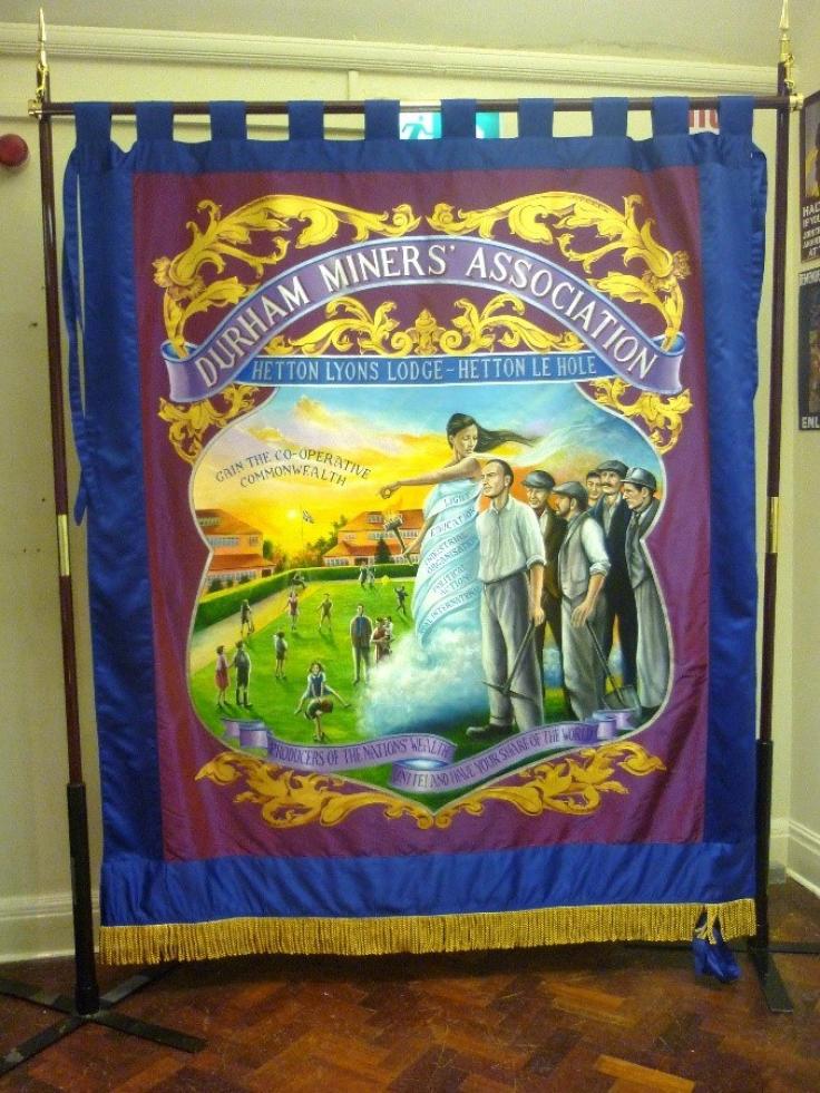 A traditional pole-mounted Miners' banner with 'Durham Miners' Association' in a scroll, under which a group of miners are following a female figure pointing the way.