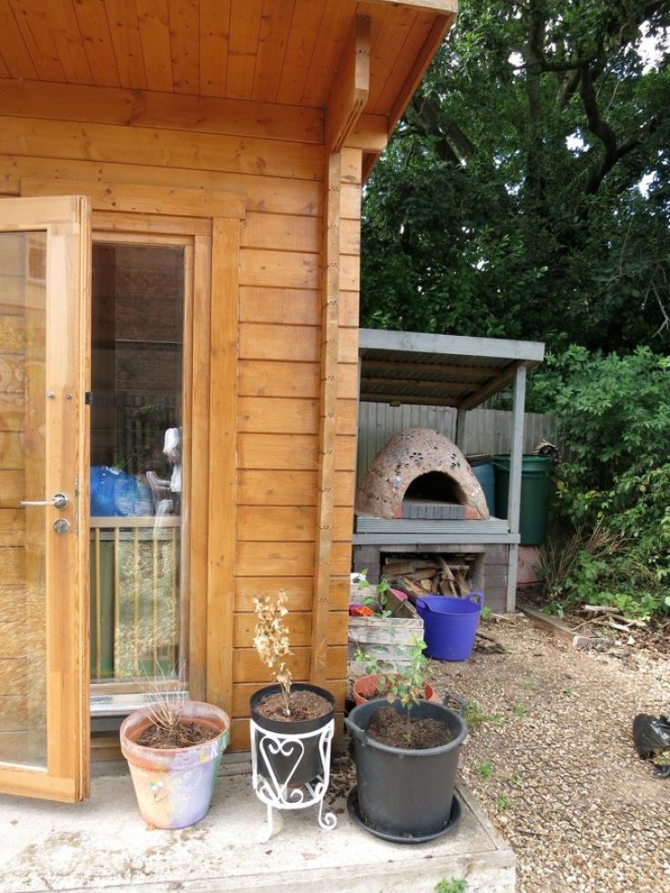 A wooden building and an outdoor clay oven with trees in the background.