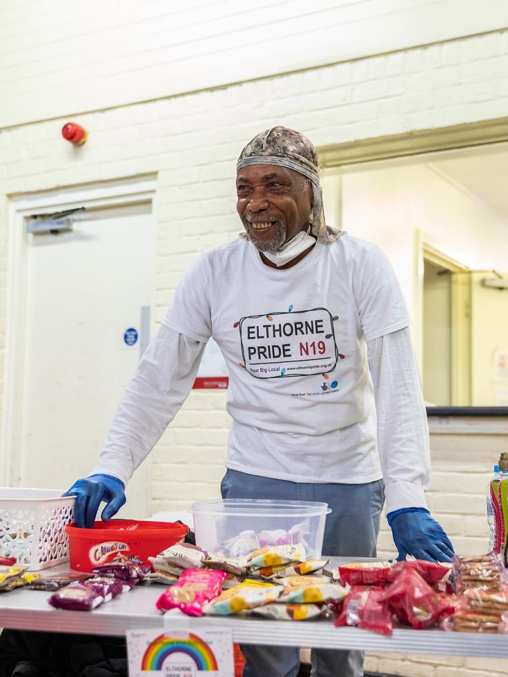 A man wears an apron reading 'I heart Elthorne Pride N19' with food on the table.