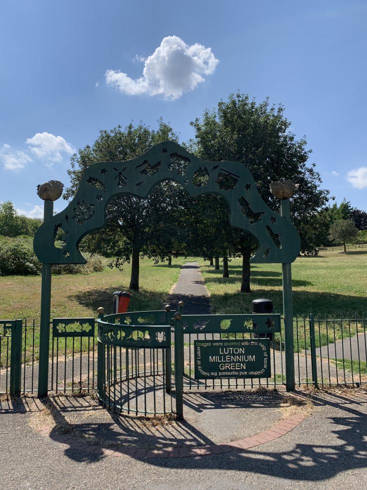 A tree-lined avenue inside green railings on which is a sign saying 'Luton Millennium Green'.