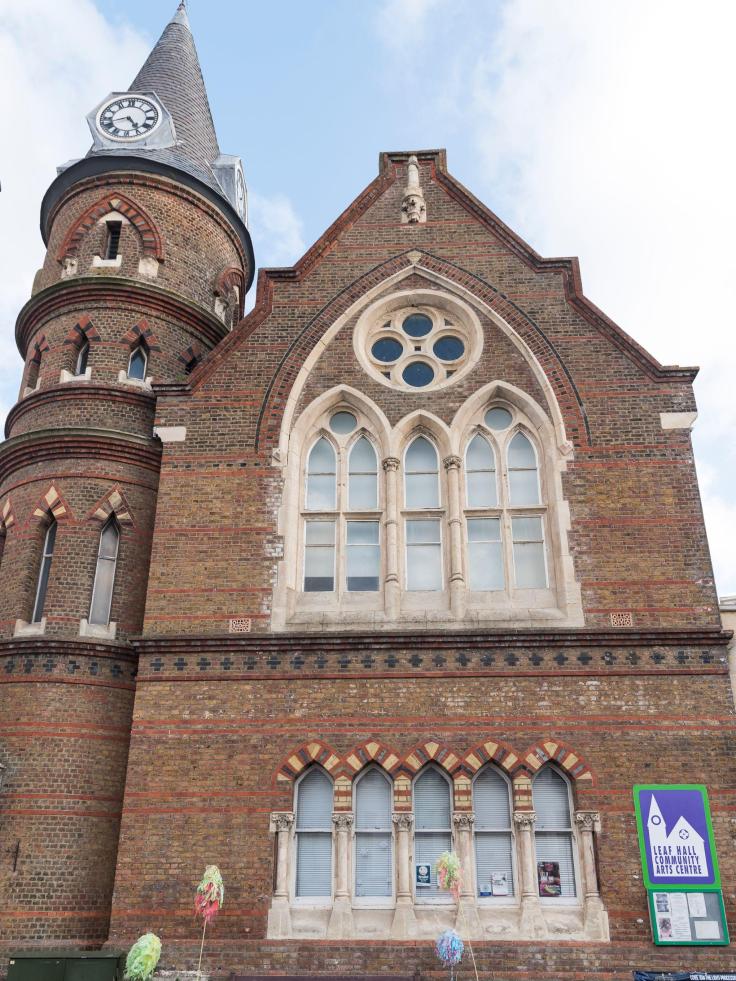 A tall red brick Victorian style building with arched windows and a turret, with a sign saying 'Leaf Hall Community Arts Centre.'